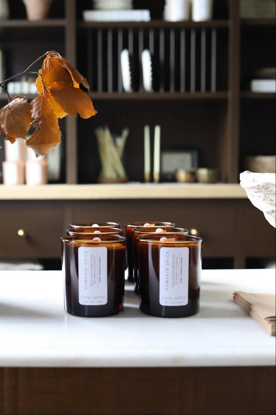 Three candles with labels on a surface, with a blurred background of shelves and decor.